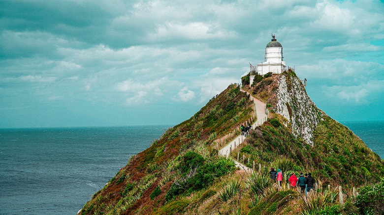 paved path climbing up to lighthouse on a hill overlooking a blue sea