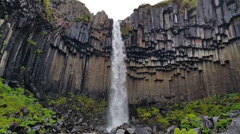 Thin waterfall falling over grey basalt pillars that seem to be suspended