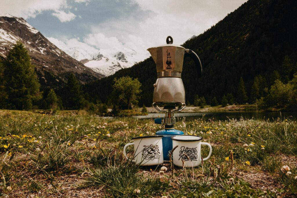 two cups and moka pot on backpacking stove in front of mountain lake